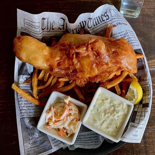 Golden-brown fish and chips with coleslaw, tartar sauce, and a lemon wedge on newspaper-print paper.