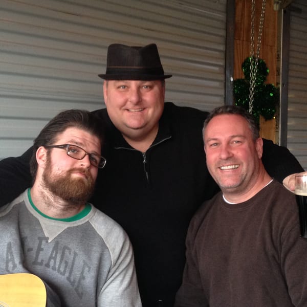 Three men smiling, one with a fedora, another with a beard and guitar, at Trooper Thorn's.