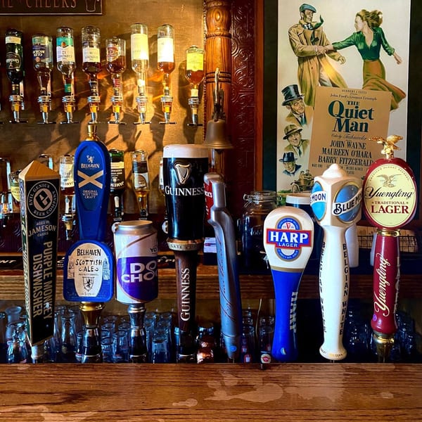 A bar tap with various beer handles, including Guinness, Harp, and Yuengling, with whiskey bottles above.