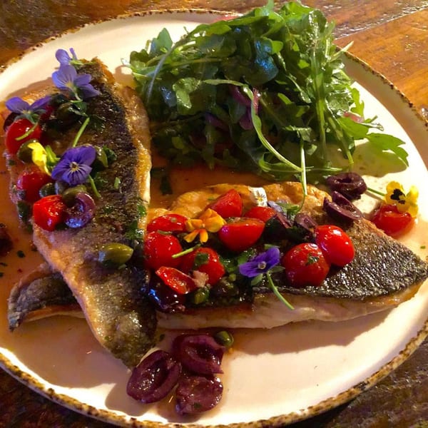 Pan-seared fish fillets with cherry tomatoes, olives, capers, edible flowers, and a side of arugula.