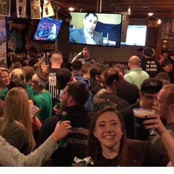 A crowded Irish pub with patrons holding drinks, watching TV screens, and socializing.