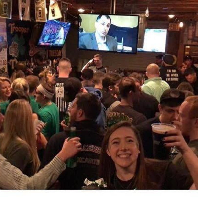A crowded Irish pub with patrons holding drinks, watching TV screens, and socializing.