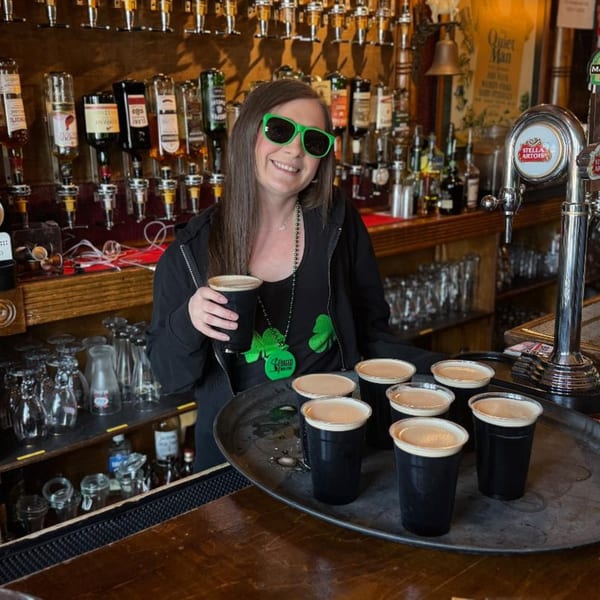 Smiling bartender in green sunglasses and shamrock beads holding a stout, with a tray of stouts.