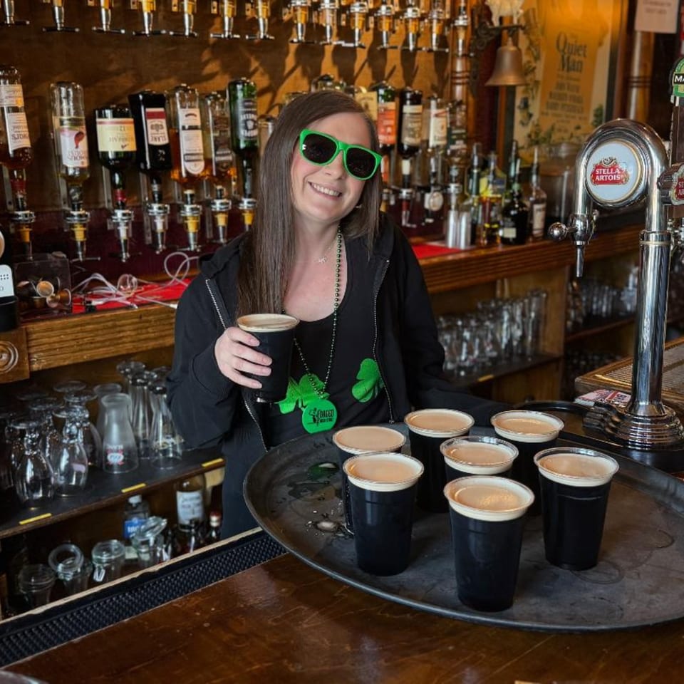 Smiling bartender in green sunglasses and shamrock beads holding a stout, with a tray of stouts.
