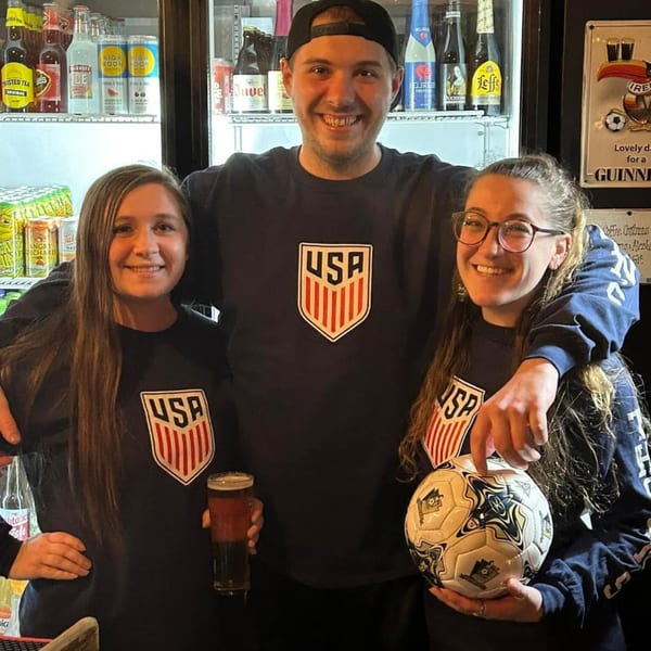 Three smiling people in USA shirts, one holding a beer, another a soccer ball, in an Irish pub.