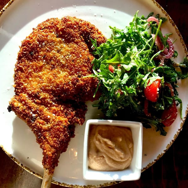 Crispy breaded pork chop with a side of fresh arugula salad and a small dish of sauce.