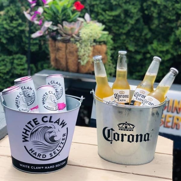 Two buckets of beverages, one with White Claw seltzers and one with Corona beers, on a wooden table.