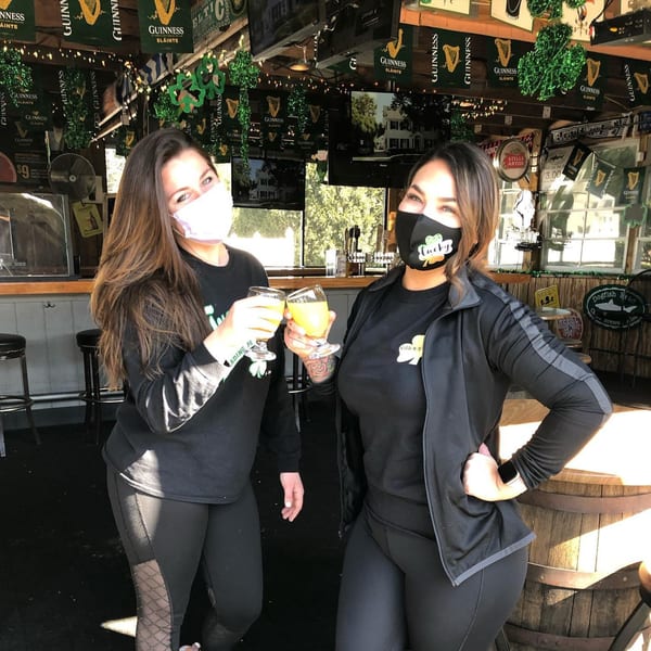 Two women in masks toast with drinks inside Trooper Thorn's Irish Beef House, decorated with Guinness banners.
