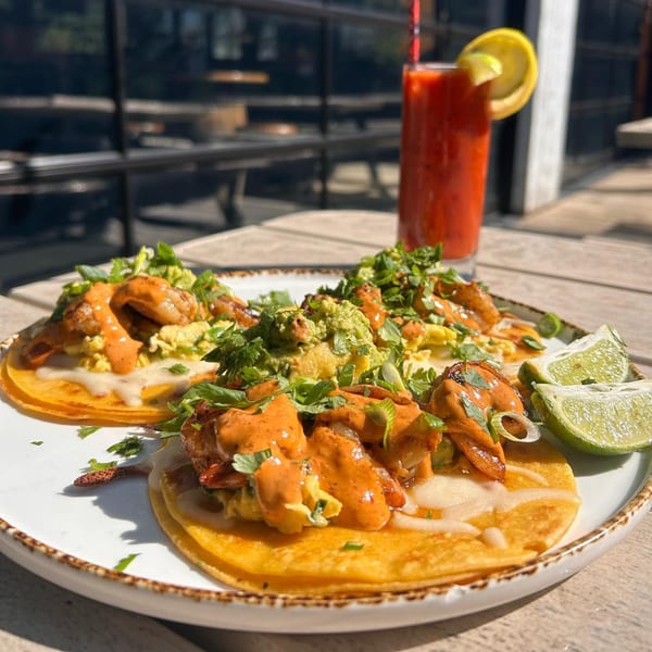 Three shrimp tacos with guacamole, sauce, and cilantro, with a Bloody Mary and lime wedges.