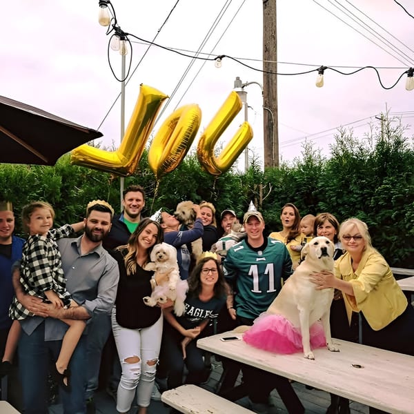 A large group of people and dogs, including a dog in a pink tutu, pose outdoors.
